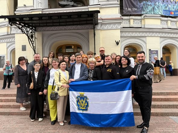 Групова фотографія делегації на сходах біля головного входу до Національної опери України. Близько двадцяти людей — чоловіків, жінок та підлітків — позують для знімка під старовинним козирком театру. У центрі на передньому плані вони тримають розгорнутий великий синьо-біло-синій прапор міста Херсона з офіційним гербом громади. Чоловік праворуч одягнений у чорну вишиванку з білим орнаментом, інші учасники — у повсякденному та класичному одязі. На задньому плані видно світлу фасадну архітектуру театру та перехожих.