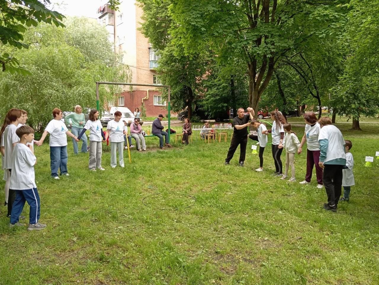 Спортивні розваги та естафети для дітей та дорослих на зеленій галявині у дворі будинку. Учасники заходу, переважно у білих футболках, вишикувалися у дві лінії та тримаються за руки. По центру чоловік у чорному одязі допомагає дівчинці пройти крізь зелений пластиковий обруч (хулахуп), не розриваючи рук. На задньому плані на лавках сидять глядачі, видно цегляні багатоповерхові будинки.