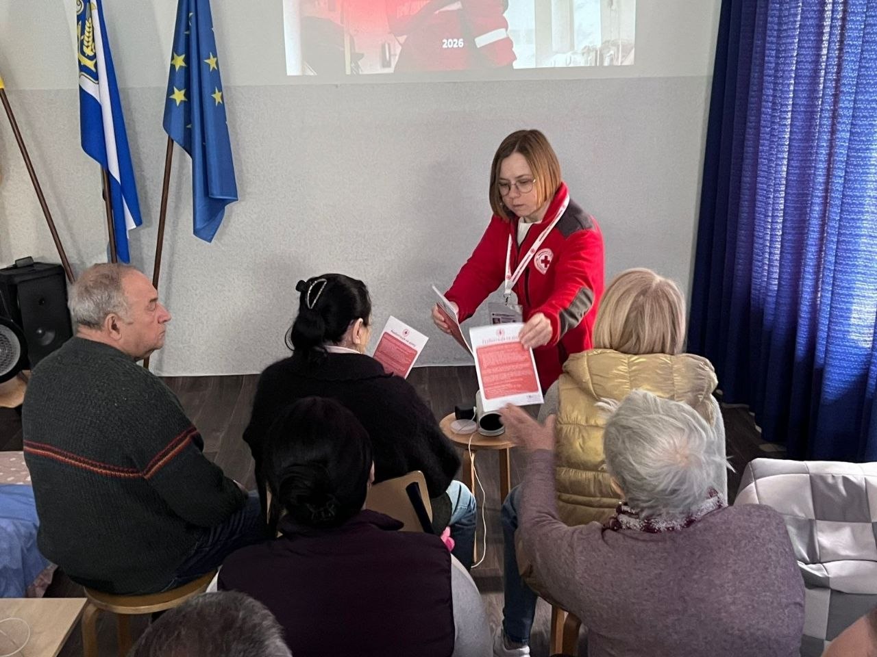 Співробітниця Червоного Хреста у фірмовій червоній куртці з логотипом організації роздає друковані інформаційні пам'ятки та буклети людям похилого віку під час зустрічі у приміщенні хабу. Люди сидять на стільцях півколом на тлі світлої стіни, де встановлено прапори України та Європейського Союзу.