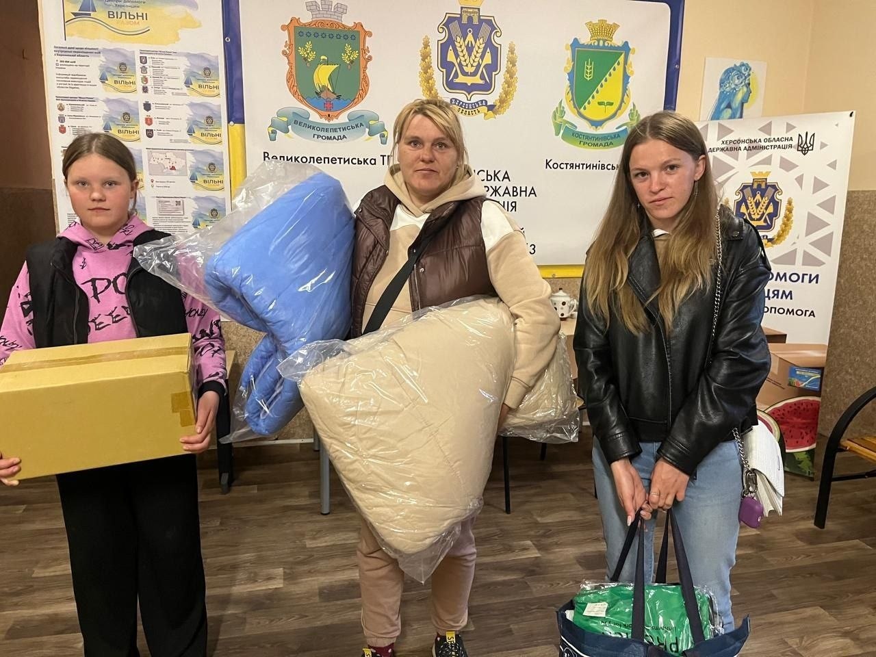 Три дівчини-переселенки позують у гуманітарному хабі з отриманою допомогою. Одна з них тримає велику картонну коробку, інша — ковдри та подушки у прозорому пластиковому пакуванні, а третя — велику дорожню сумку. Вони стоять перед білим банером, на якому зображені герби Великолепетиської та Костянтинівської територіальних громад Херсонської області.