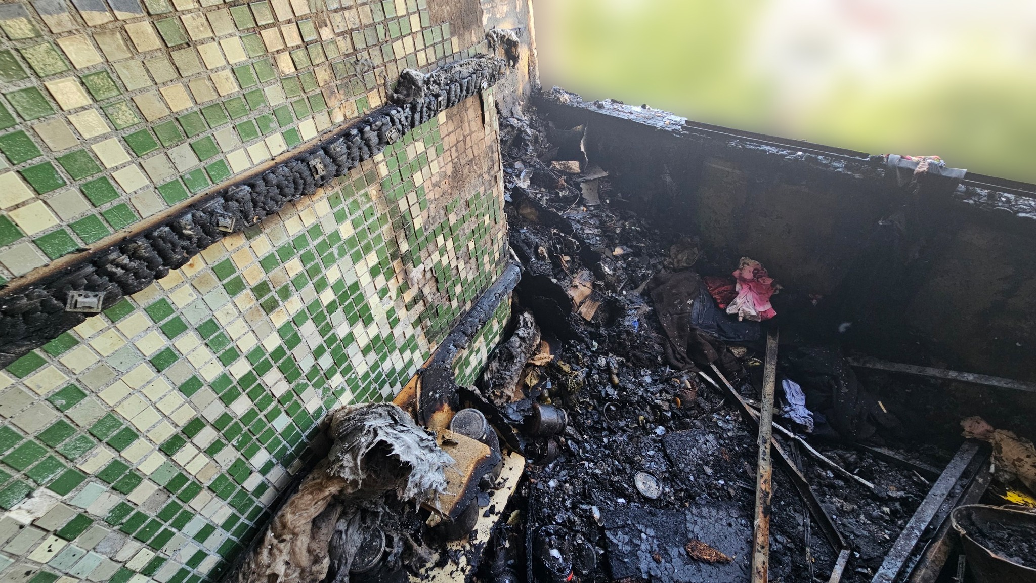 Зруйнований та випалений вщент балкон багатоповерхівки, вид згори. На підлозі лежить суцільний шар попелу, вугілля, уламків будматеріалів та згорілих побутових речей. Ліворуч видно уцілілу стіну, оздоблену дрібною квадратною плиткою зеленого та бежевого кольорів, на якій залишився горизонтальний слід від кіптяви. Тло за межами балкона розмите.