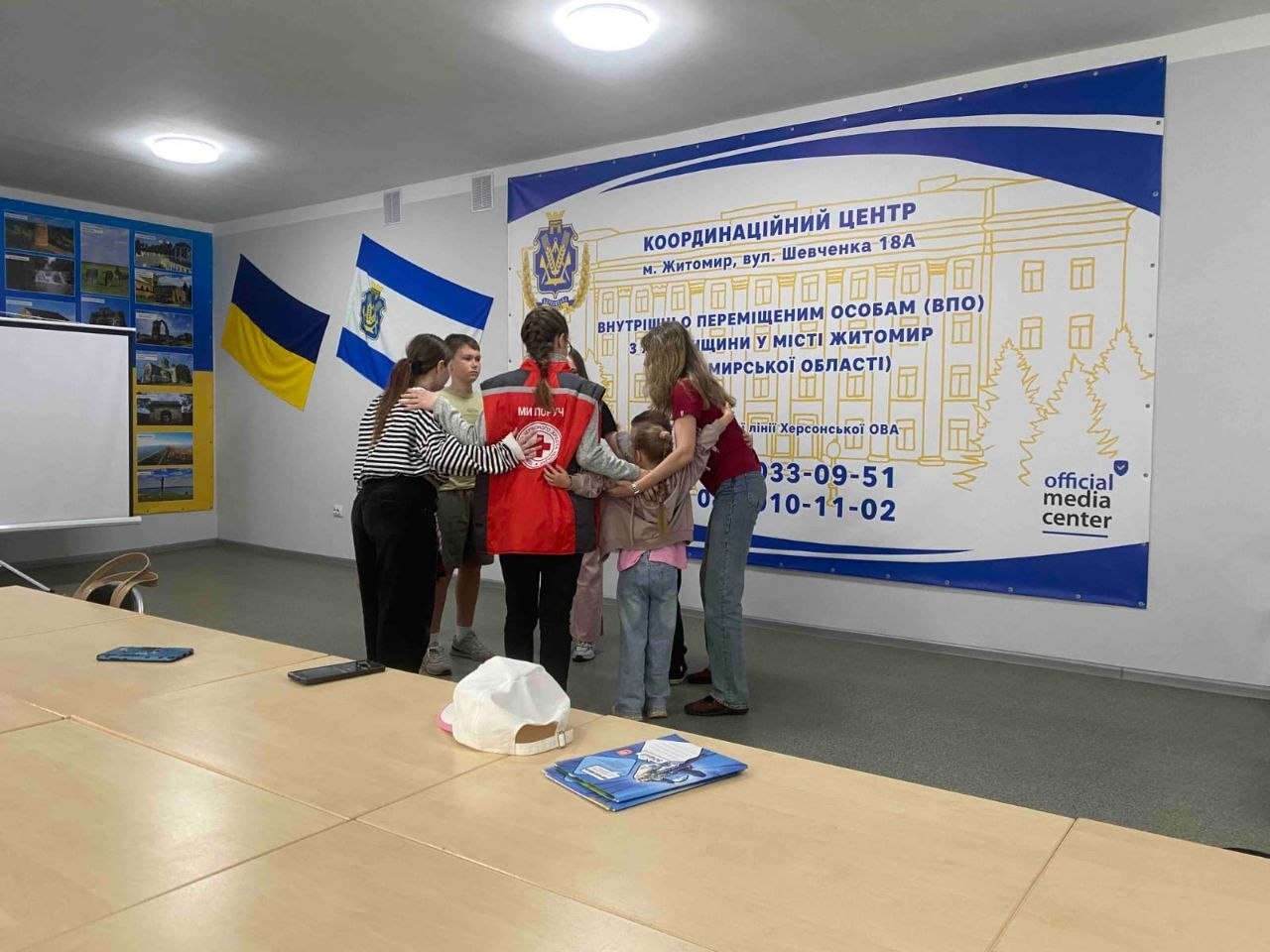 Волонтери та діти стоять колом, обнявшись за плечі, під час інтерактивного заходу в офіційному центрі підтримки. Серед дорослих — дівчина у червоно-сірому жилеті з емблемою Червоного Хреста. На задньому плані розташований великий біло-синій банер із написом «КООРДИНАЦІЙНИЙ ЦЕНТР м. Житомир, вул. Шевченка 18А внутрішньо переміщеним особам (ВПО) з Херсонщини у місті Житомир (Житомирської області)» та телефонами гарячої лінії. На стіні ліворуч висять прапори України та міста Херсона.