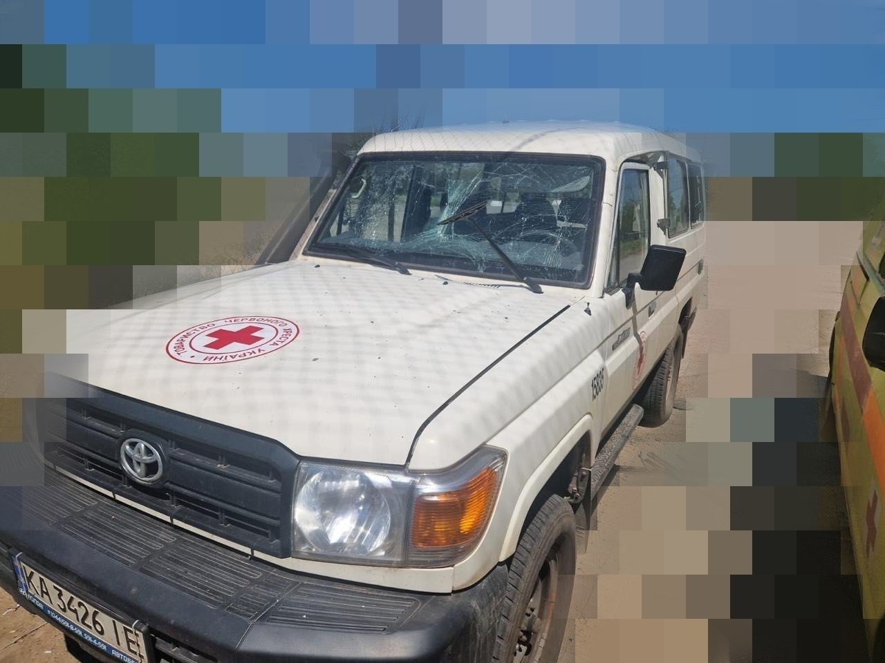 Білий позашляховик Toyota Land Cruiser із пошкодженнями від обстрілу або вибухової хвилі. Лобове скло автомобіля повністю вкрите густою сіткою глибоких тріщин та має осипання. На капоті нанесено кругле червоно-біле маркування Товариства Червоного Хреста України. Автомобіль має київські номери «KA 3426 IE».
