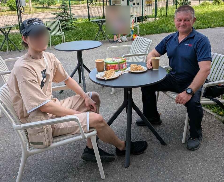 Хлопець-підліток у бежевому костюмі (шорти та футболка), чорних кросівках і кепці сидить за круглим столиком літнього майданчика кафе поруч із дорослим чоловіком у синій футболці поло. На столі перед ними стоять паперові стаканчики з кавою, тарілки зі шматочками торта та кругла коробка. Обличчя підлітка приховане за розмиттям. На задньому плані видно зелену зону, паркан та інші вуличні меблі.