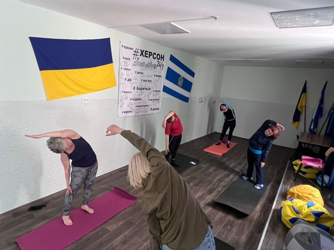 П'ять жінок різного віку займаються фітнесом або йогою у спортивній залі. Вони стоять на індивідуальних різнокольорових килимках (карематах) і синхронно виконують нахили тулуба вбік з піднятою рукою. На стіні зали закріплені прапори України та Херсона, а також мотиваційний плакат із патріотичними гаслами.
