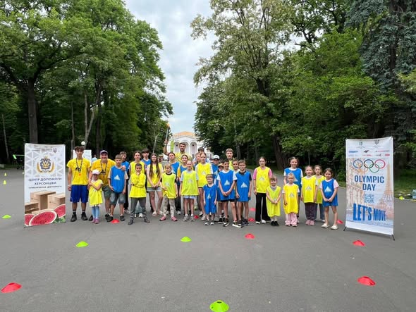 Загальне планове фото всіх учасників «Olympic Day» у парку. Понад двадцять дітей та підлітків вишикувалися в один довгий ряд по центру великого асфальтованого майданчика. З обох боків від групи встановлено тематичні банери: ліворуч — стенд «Центр допомоги херсонцям», праворуч — банер «Olympic Day. Let's Move» з олімпійськими кільцями та зображенням Ейфелевої вежі. На передньому плані на асфальті півколом розставлені червоні та зелені маркувальні фішки. На задньому плані розкинувся густий зелений парк із білою будівлею вдалині.