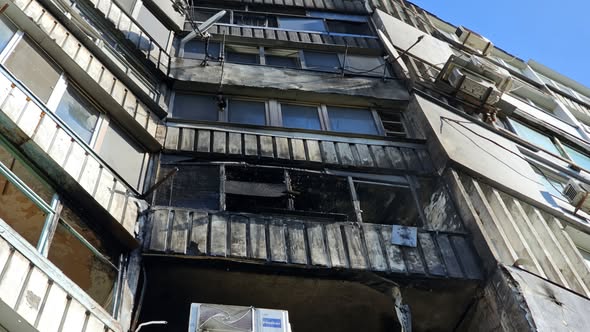 Вид знизу під гострим кутом на пошкоджений обстрілом фасад житлового будинку. Вогонь повністю знищив скління одного з балконів, перетворивши його на обвуглену бетонну нішу. Навколо зруйнованої конструкції чітко видно зони задимлення, кіптяву на залізобетонних панелях та пошкоджені рами сусідніх квартир на тлі чистого синього неба.