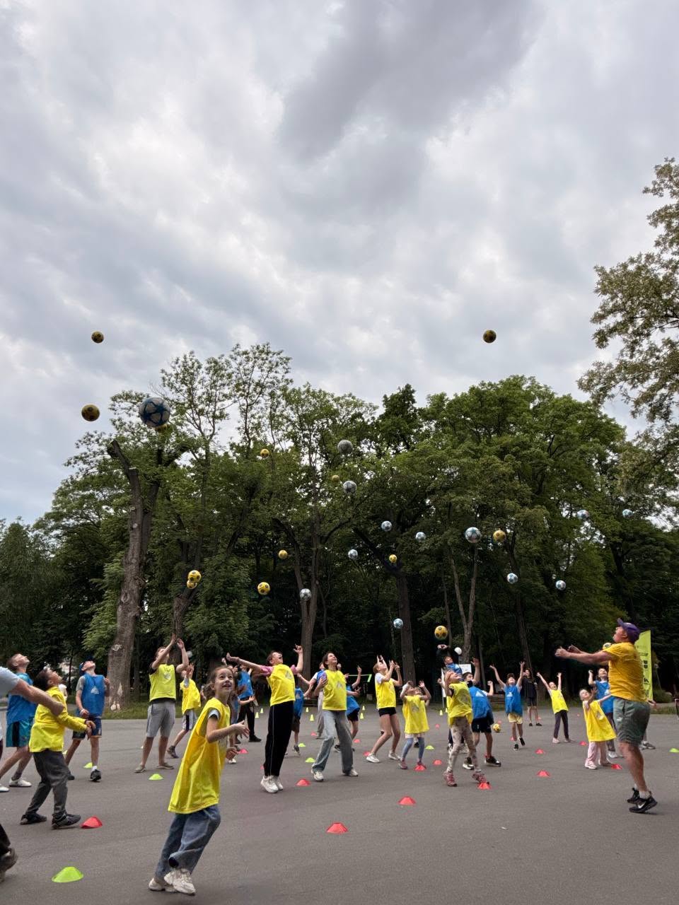 Момент виконання спортивної вправи під відкритим хмарним небом. Група дітей та підлітків у синіх і жовтих маніжках одночасно підкинули вгору велику кількість різнокольорових м'ячів (футбольних, волейбольних і малих тенісних). Усі учасники стоять на майданчику, піднявши голови та руки вгору, спостерігаючи за польотом м'ячів. Справа тренуванням керує чоловік у жовтій футболці та фіолетовій кепці. Позаду дітей височіють великі зелені дерева.