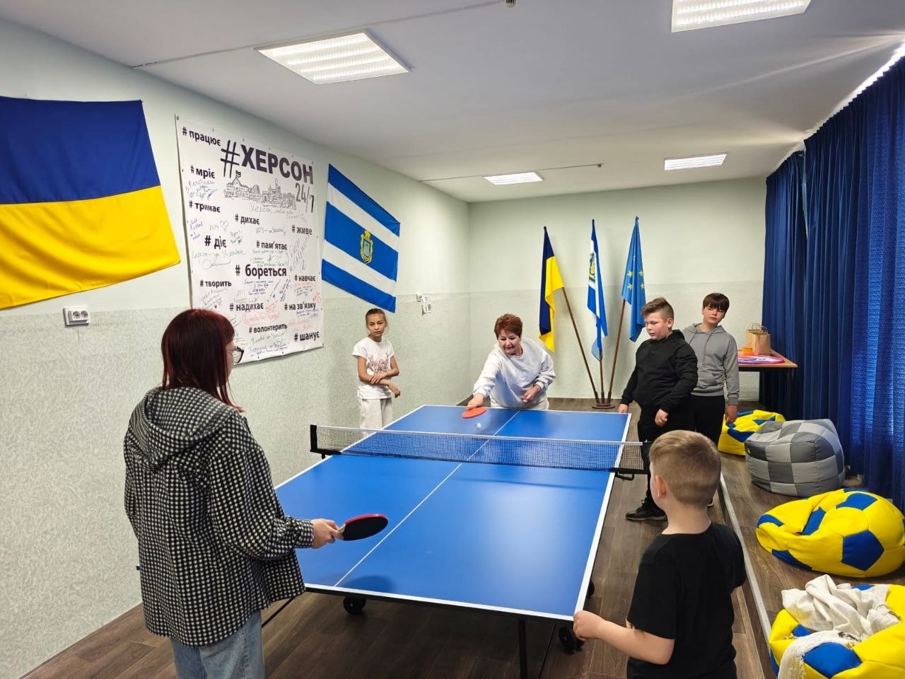 Гра в настільний теніс в ігровій кімнаті гуманітарного центру. Жінка похилого віку та дівчина з червоним волоссям грають за синім тенісним столом. Поруч стоять і спостерігають за грою троє дітей. На світлій стіні кімнати висить державний прапор України, синьо-білий прапор міста Херсона та великий плакат із написами «ХЕРСОН 24/7» та патріотичними хештегами.