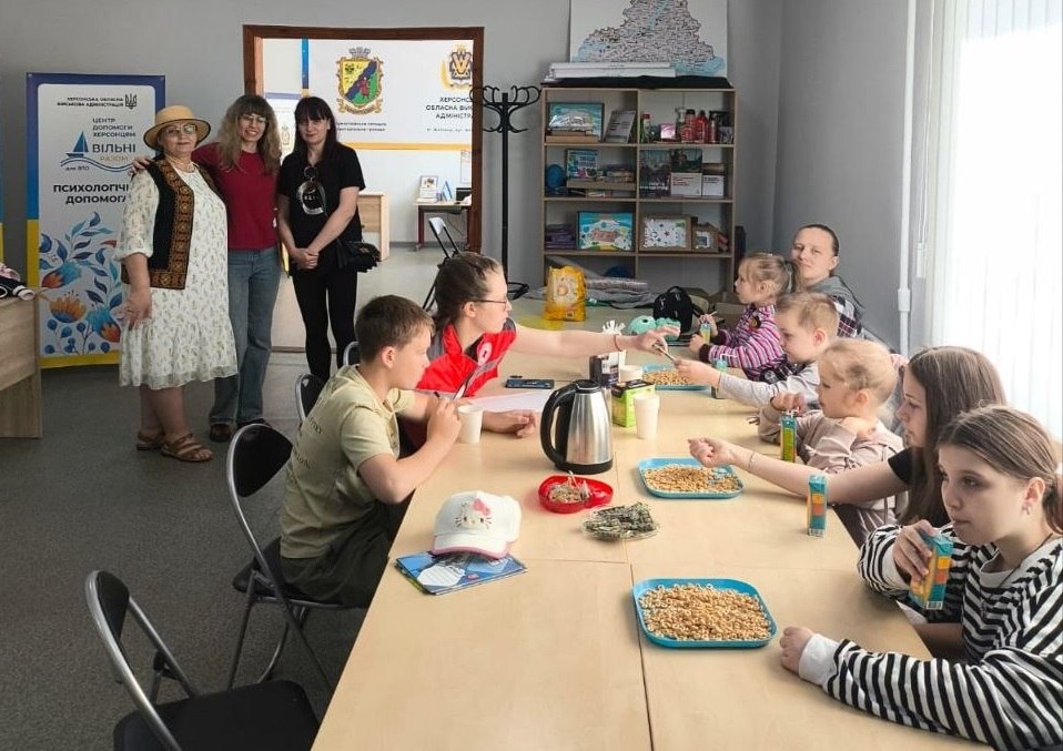 Група дітей та матерів сидить за довгим світлим столом у кімнаті підтримки. Вони розмовляють, п'ють сік і пригощаються солодощами. По центру столу стоїть металевий електрочайник. Волонтерка у фірмовому жилеті Червоного Хреста передає щось дітям. Ліворуч на задньому плані стоять три жінки, поруч із ними — банер «Центр допомоги херсонцям "Вільні разом"» з анонсом психологічної допомоги. У дверному отворі видно герб Житомирської міської громади.