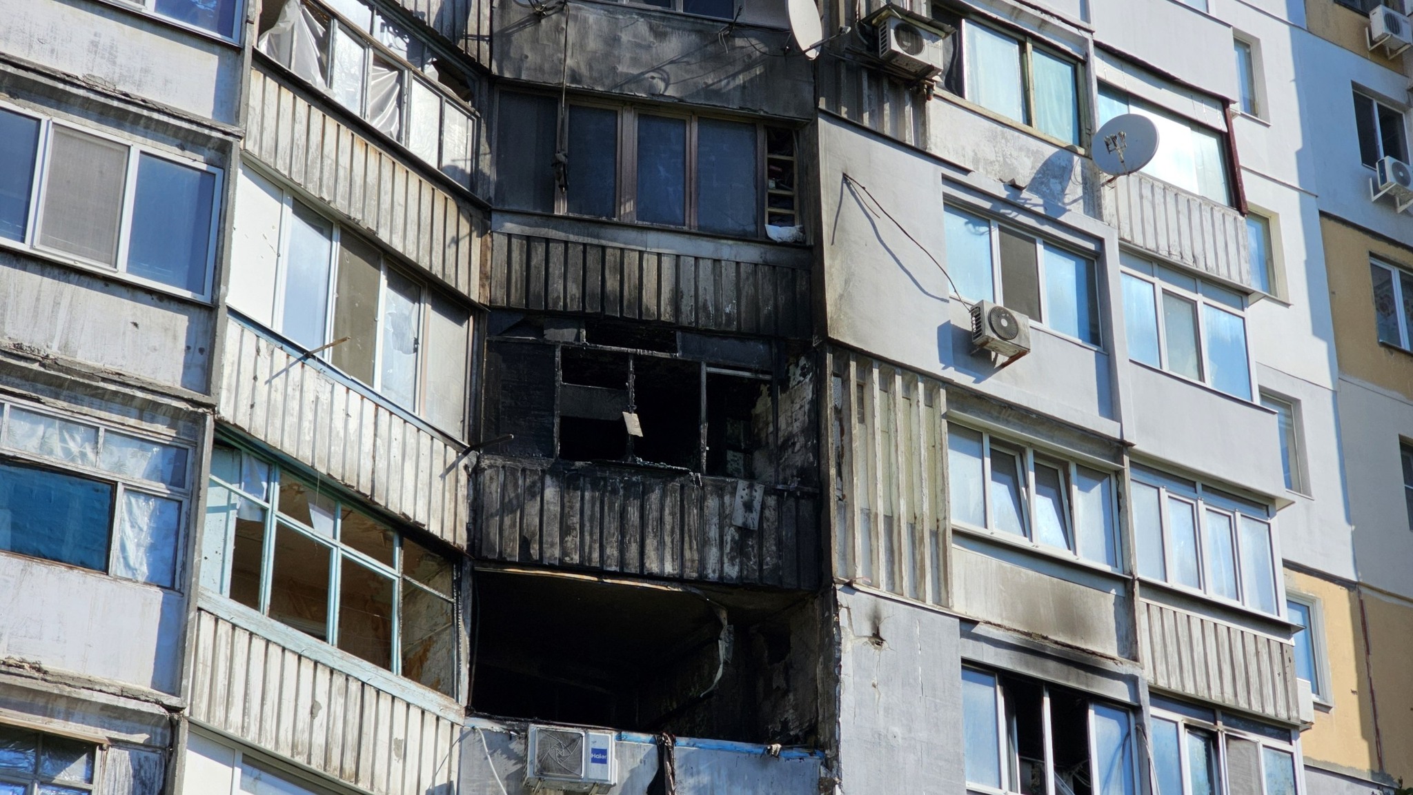 Фрагмент фасаду панельного багатоповерхового будинку з наслідками прямого влучання. Один із центральних балконів повністю випалений, його віконні рами знищені, а внутрішній простір зяє чорною пусткою. Сусідні плити фасаду, блоки кондиціонерів та вікна вище і нижче епіцентру вибуху покриті сильними чорними слідами від пожежі та диму.