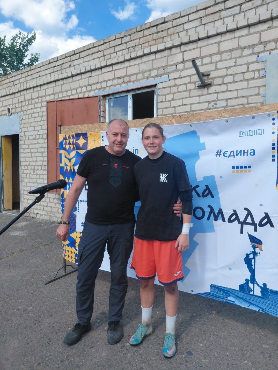 Чоловік у чорній футболці з вишитим орнаментом та дівчина-арбітр у чорній спортивній кофті й помаранчевих шортах позують разом на тлі цегляної будівлі. Вони стоять поруч перед білим інформаційним банером із написом «#єдина громада». Ліворуч на передньому плані видно стійку з мікрофоном.