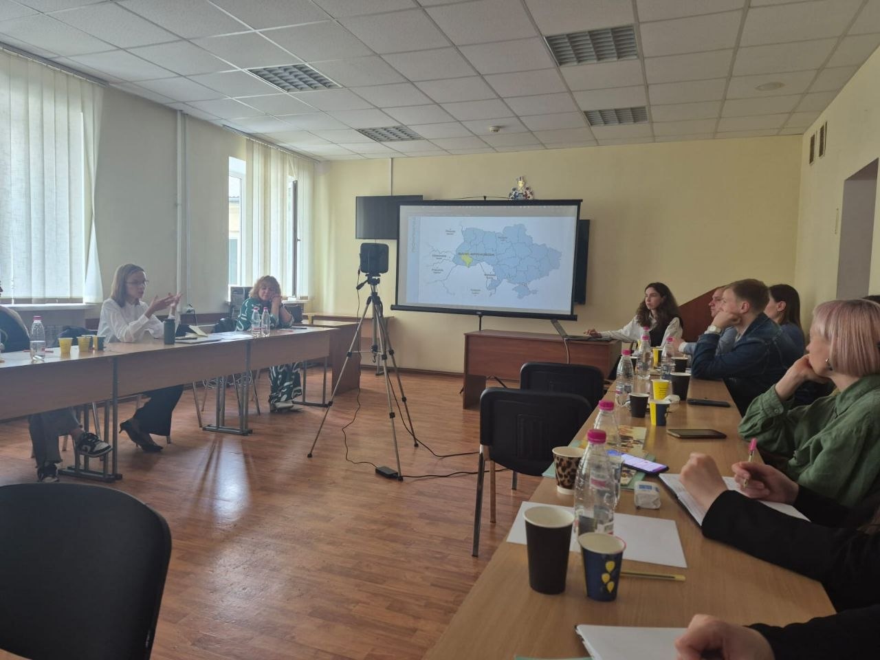 Робоче засідання або семінарія в навчальній аудиторії під час конгресу. Зліва за довгим столом сидять лекторки; одна з них, у білій блузі, говорить і жестикулює. Навпроти них, праворуч, за партами сидять слухачі (молоді люди, дівчата), які роблять записи у зошитах та ноутбуках. На столах стоять пляшки з водою та паперові стаканчики. На задньому плані встановлено великий проєкційний екран, на якому демонструється контурна карта України з виділеною жовтим кольором Хмельницькою областю. Поруч на штативі закріплена відеокамера.