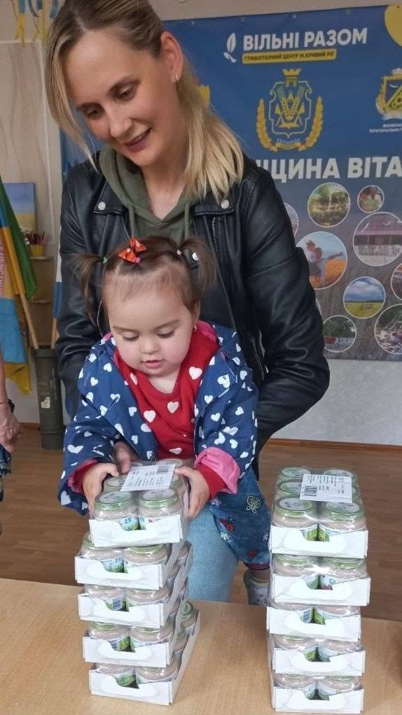 Молода жінка у чорній шкіряній куртці допомагає маленькій дівчинці тримати скляні баночки з дитячим харчуванням. Дівчинка з двома хвостиками одягнена у синю курточку в білі сердечка та червону кофту. Перед ними на дерев'яному столі вибудувані у дві високі стопки блоки з баночками дитячого пюре в білих картонних підставках. На задньому плані висить синій фірмовий банер гуманітарного центру «Вільні разом».