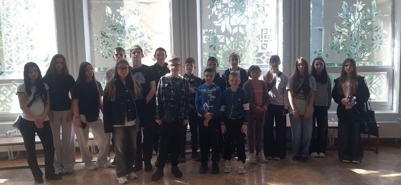 Групова фотографія підлітків у просторому світлому приміщенні. Близько п'ятнадцяти хлопців та дівчат шкільного віку вишикувалися в один ряд для спільного знімка. Вони одягнені у повсякденний одяг (худі, футболки, джинси, спортивні штани). Позаду них розташовані великі вікна, прикрашені білими паперовими витинанками з рослинними та геометричними візерунками. Світло з вікон створює силуетний ефект, через що обличчя учасників частково затемнені.