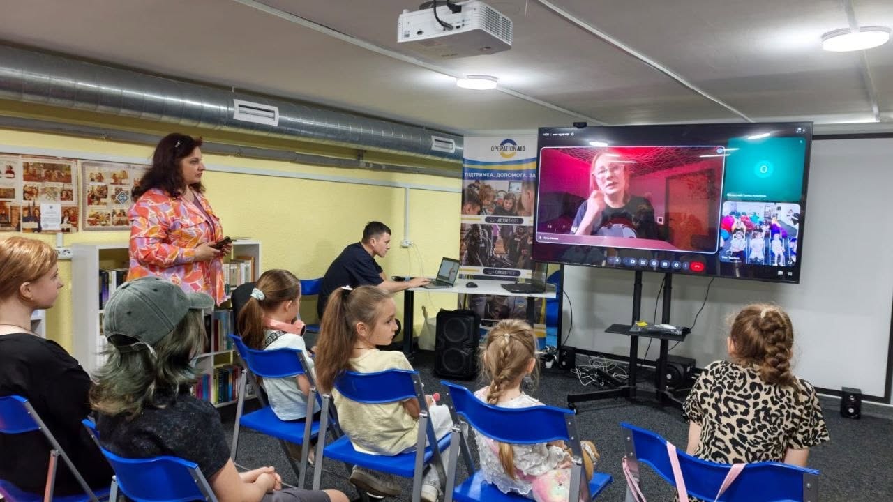 Проведення онлайн-зустрічі або лекції для дітей у затишному приміщенні з низькою стелею.  У правій частині кадру на стійці встановлено великий плаский телевізор, на екрані якого через сервіс відеозв'язку транслюється зображення спікерки — жінки в окулярах на тлі кімнати з рожевим підсвічуванням. У правому кутку монітора видно додаткові вікна інтерфейсу конференції з іншими учасниками.  Перед екраном півколом на синіх розкладних стільцях сидять слухачі — переважно діти та підлітки, повернуті спиною до камери. Серед них виділяється дівчинка в кепці ліворуч та дівчинка в блузці з леопардовим принтом праворуч.  У лівій частині приміщення стоїть жінка у яскравій помаранчево-рожевій сорочці з візерунками, яка тримає в руках телефон і спостерігає за процесом. За нею, біля стіни, розташовані книжкові полиці та плакати. По центру, на задньому плані, за столом із ноутбуком сидить молодий чоловік у чорній футболці, який забезпечує технічний супровід трансляції. Поруч із ним встановлено інформаційний стенд-ролап із логотипом благодійної організації «OPERATION AID». На стелі закріплено увімкнений білий проектор та вентиляційні труби.