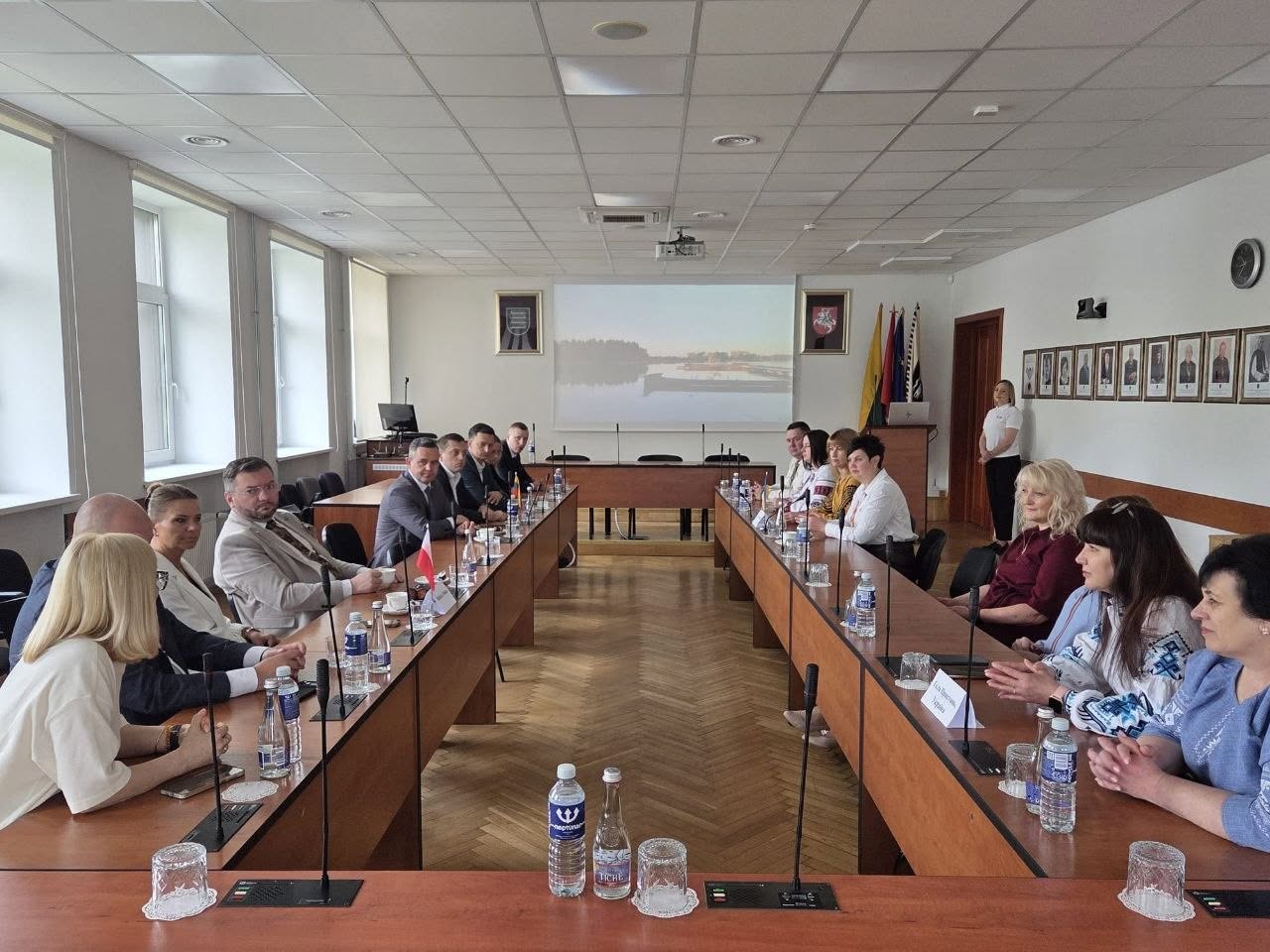 Офіційне засідання делегацій у конференц-залі. За великим довгим столом у формі літери «U» одне навпроти одного сидять учасники зустрічі — чоловіки в ділових костюмах та жінки, частина з яких вдягнена у вишиванки. На столі перед ними розставлені пляшки з водою, склянки, мікрофони та встановлено маленький прапорець Польщі. На стіні в кінці залу працює проектор, що транслює зображення річкового пейзажу. Справа біля трибуни стоїть дівчина у білій футболці. На правій стіні в ряд розвішані офіційні портрети у рамках.