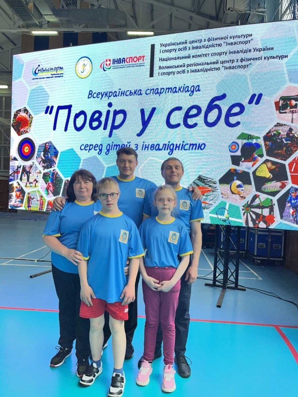 Групова фотографія п'ятьох людей у спортивному залі на тлі великого світлодіодного екрана спартакіади. На передньому плані стоять двоє дітей: хлопчик в окулярах та червоних шортах і дівчинка в рожевих спортивних штанях. Обидва одягнені в однакові яскраво-блакитні футболки з жовтою облямівкою та невеликим гербом. За ними стоять троє дорослих (двоє чоловіків та жінка) у такому ж фірмовому блакитному одязі. На великому екрані позаду них нанесено логотипи системи «Інваспорт» та великий напис: «Всеукраїнська спартакіада “Повір у себе” серед дітей з інвалідністю».