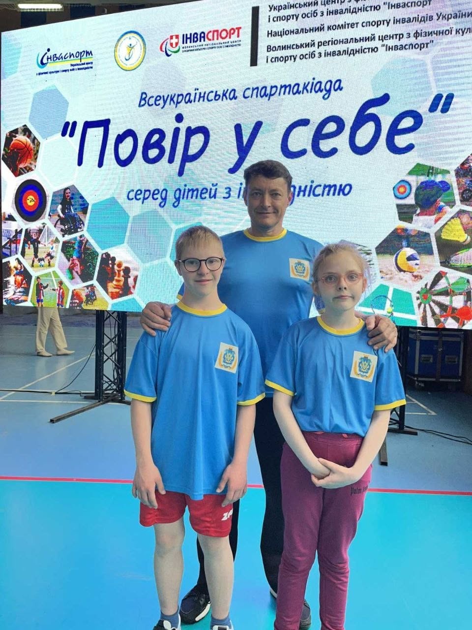 Фотографія великим планом у приміщенні спорткомплексу. Двоє юних учасників спартакіади (хлопчик і дівчинка в окулярах) стоять поруч перед камерою, одягнені у блакитні фірмові футболки з жовтими елементами. Позаду них по центру стоїть чоловік-тренер, який поклав руки їм на плечі. На задньому плані працює великий цифровий банер із символікою «Інваспорт» та назвою заходу «Повір у себе». Ліворуч на розмитому тлі видно ноги іншої людини, що проходить повз.
