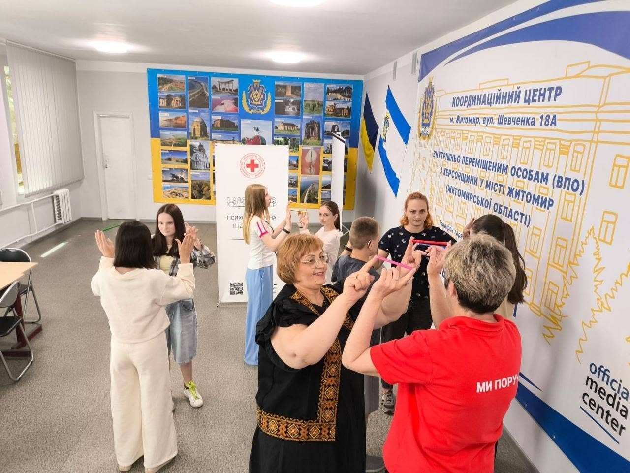 Процес проведення групового заходу у просторому кабінеті гуманітарного центру. На передньому плані праворуч зображено спиною до камери жінку в яскравій червоній футболці з написом «МИ ПОРУЧ» та чорних штанах, яка рухається кімнатою. Лівіше від неї повз камеру проходить жінка у темній футболці з дрібним візерунком та сірих джинсах. У центрі кадру на розкладних чорних стільцях сидять діти та підлітки.  На синьо-жовтому тлі далекої стіни розміщено великий стенд із мініатюрними фотографічними краєвидами Херсонщини, перед яким стоїть ролап із червоним хрестом. Справа на стіні закріплено великий інформаційний банер Координаційного центру допомоги ВПО з Херсонщини у місті Житомир із телефонами гарячої лінії. Поруч висять державний прапор України та синьо-білий прапор Херсона.