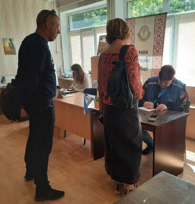 Робочий процес реєстрації відвідувачів у державній установі чи гуманітарному центрі. Справа за столом сидить співробітниця у фірмовому синьому жилеті поверх квітчастої кофти і заповнює журнал. Перед її столом стоїть жінка у довгій темній спідниці та блузці з відкритими плечима та рюкзаком. Лівіше стоїть чоловік у чорному светрі та темних штанах, спостерігаючи за процесом. На задньому плані за іншим столом видно ще одну співробітницю за комп'ютером та банер із гаслом «Разом робимо більше!».