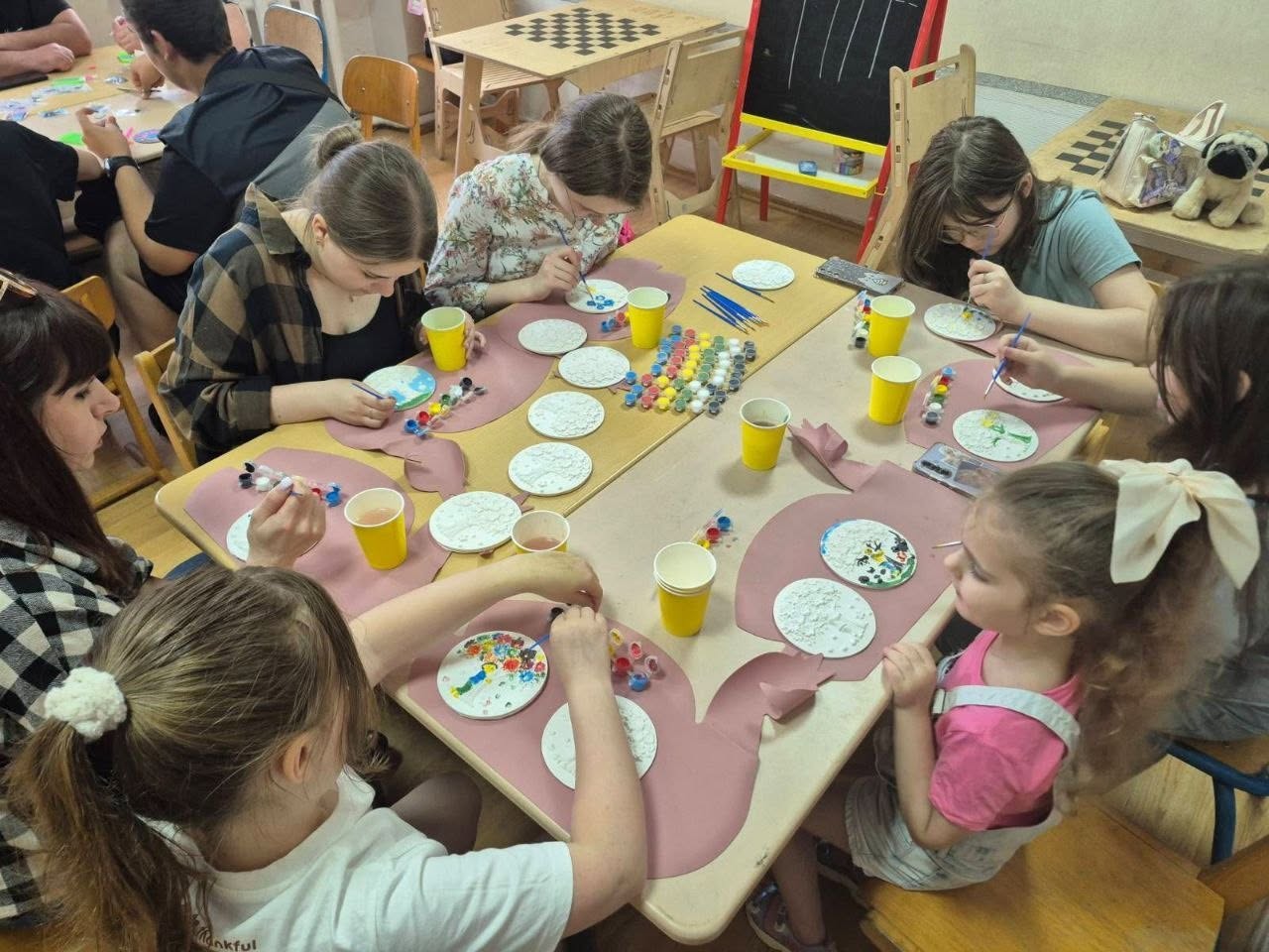 Вид згори на робочий стіл під час майстер-класу, де шестеро дівчат різного віку розписують круглі гіпсові чи дерев'яні рельєфні заготовки. На передньому плані зліва дівчинка з русявим волоссям у білій футболці розфарбовує візерунок яскравими фарбами. Праворуч від неї сидить маленька дівчинка у рожевій майці та світлому комбінезоні з великим бежевим бантом у волоссі. На столі, застеленому рожевим захисним папером, розкладено заготовки з різними стадіями готовності малюнків, палітри, набори акрилових фарб та жовті стаканчики для води. На задньому плані видно м'яку іграшку мопса.