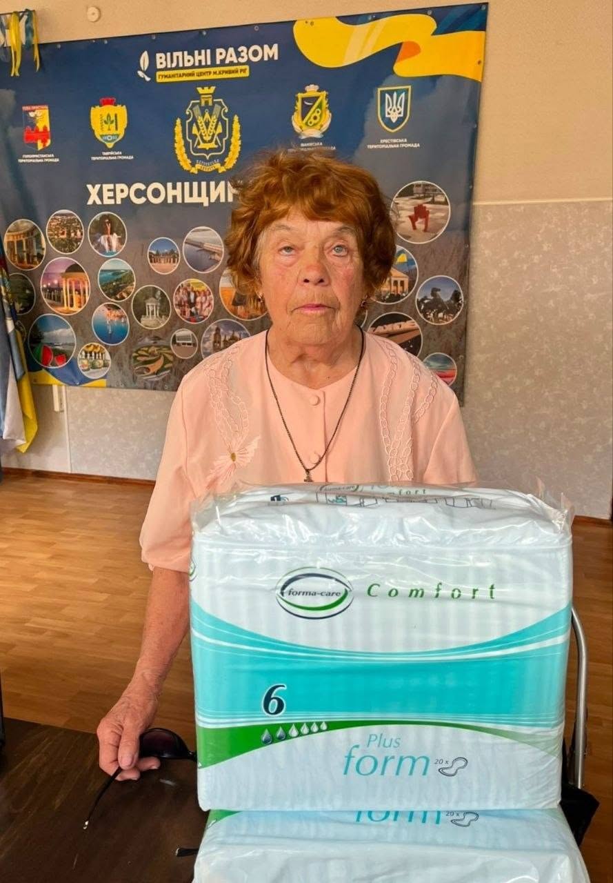 Літня жінка з рудим коротким волоссям, одягнена у світло-рожеву блузку, стоїть біля столу в приміщенні хабу. Вона обома руками тримає велику прозору упаковку з гігієнічними підгузками для дорослих фірми «Forma-Care Comfort, 6 Plus». Жінка дивиться повз камеру. Безпосередньо за її спиною розташований синій банер із символікою Херсонської області та круглими фотоілюстраціями регіону.