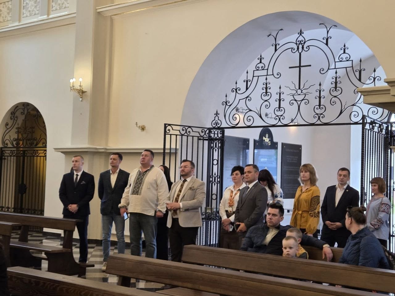 Група людей під час перебування всередині собору або костелу. Чоловіки та жінки стоять уздовж проходу між дерев'яними лавами для прихожан, повернувши голови в один бік і зосереджено спостерігаючи за подією. Один із чоловіків одягнений у світлу вишиванку, інші — в офіційні піджаки чи сорочки. На передньому плані на лаві сидить чоловік із маленькою дитиною. На задньому плані видно високу білу арку та декоративну чорну металеву перегородку з кованим візерунком, релігійним хрестом і пам'ятними табличками.