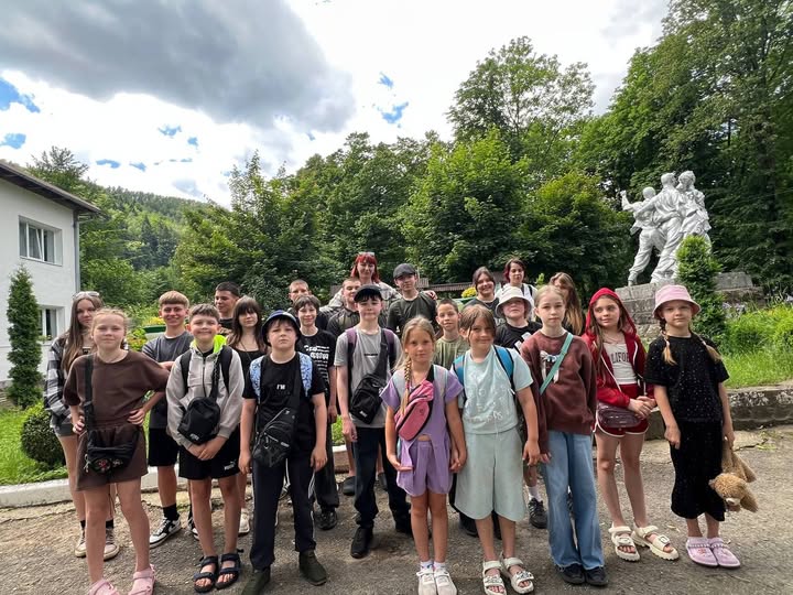 Групова фотографія дітей та підлітків на екскурсії або під час відпочинку. Близько двадцяти учасників різного віку вишикувалися у два ряди на асфальтованому майданчику. Діти одягнені у літній повсякденний одяг (футболки, шорти, кофти, панами), у багатьох із них через плече надіті маленькі спортивні сумки-бананки.  На передньому плані по центру стоять дві дівчинки: одна в бузковому костюмі з рожевою сумкою, інша — у світло-м'ятному вбранні. Праворуч стоїть дівчинка в рожевій панамі, яка тримає під пахвою м'яку іграшку ведмедика. Ліворуч на передньому плані позує дівчинка у коричневому костюмі, спершись рукою на бік.  На задньому плані зліва видно частину двоповерхової білої будівлі з вікнами. Праворуч височіє великий пам'ятник сріблястого кольору, що зображує групу людей. Навколо майданчика ростуть густі зелені дерева та чагарники, а вгорі розкинулося хмарне небо з пролісками синяви.