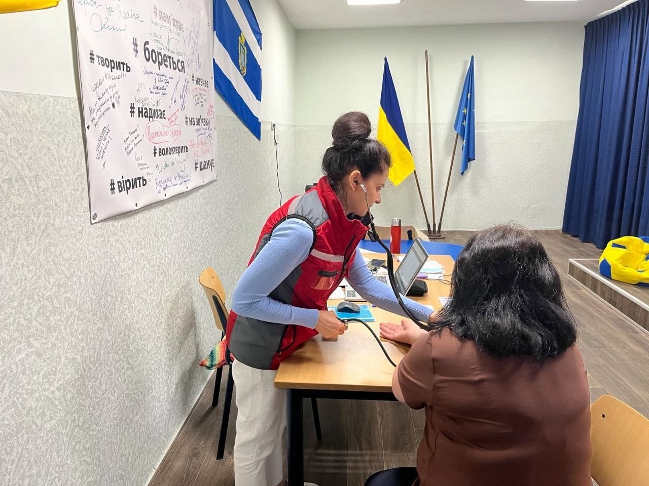 Процес вимірювання артеріального тиску пацієнтці в кабінеті хабу. Співробітниця медичної чи волонтерської організації у червоно-сірому жилеті поверх блакитної кофти стоїть біля столу, використовуючи механічний тонометр та стетоскоп. Навпроти неї спиною до камери сидить жінка у коричневій футболці, яка поклала ліву руку на стіл для процедури. На дерев'яному столі також стоїть відкритий ноутбук, лежать документи та комп'ютерна миша. На задньому плані видно прапори України та Європейського Союзу, а ліворуч — частину плаката з написами.