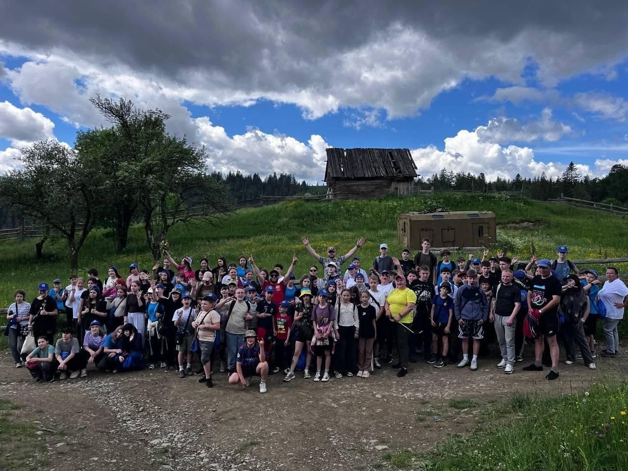 Спільне панорамне фото великої групи дітей та дорослих на гірській галявині. Кілька десятків учасників табору або екскурсії стоять у кілька рядів на ґрунтовій дорозі серед зеленої трави. Один із чоловіків у центрі підняв обидві руки вгору, вітаючи камеру. На пагорбі позаду групи стоїть стара дерев'яна хатина з напівзруйнованим дахом, а поруч із нею — залізний військовий кунг чи вагончик захисного кольору. На задньому плані розкинувся густий хвойний ліс під синім небом із купчастими хмарами.