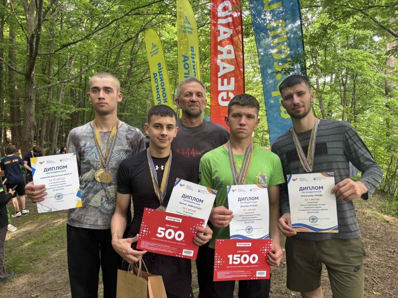 Групове фото призерів змагань з гірського туризму у лісовій місцевості. П'ятеро чоловіків стоять в один ряд на тлі високих вертикальних банерів жовтого, червоного та блакитного кольорів. Чотири молоді спортсмени тримають у руках офіційні дипломи та медалі на шиї. Двоє хлопців у центрі додатково тримають червоні подарункові сертифікати від компанії «Gorgany» номіналом «500» та «1500» гривень. Позаду них по центру стоїть старший чоловік-тренер у сірій футболці. У лівій частині кадру на задньому плані видно інших учасників змагань.