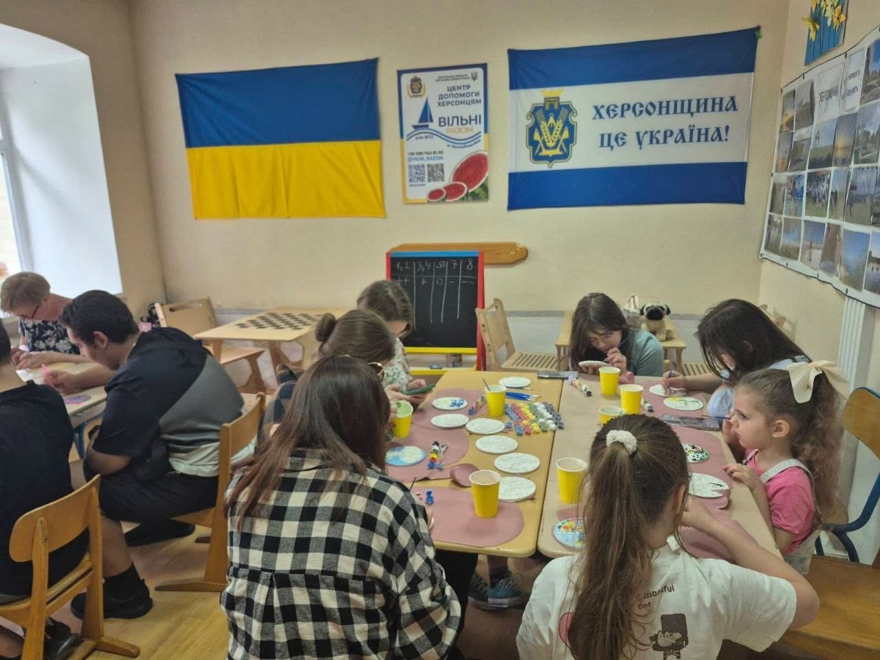 Загальний план кімнати, де близько десяти дітей та підлітків сидять за столами й займаються розписом круглих заготовок. На передньому плані спиною до камери зображено дівчину в чорно-білій сорочці в клітинку та дівчинку зі світлим волоссям, зібраним у хвіст. На столах розставлені жовті пластикові стаканчики та баночки з фарбами. На бежевій стіні на задньому плані розвішані: державний прапор України, плакат волонтерського хабу з написом «ЦЕНТР ДОПОМОГИ ХЕРСОНЦЯМ "ВІЛЬНІ РАЗОМ"» та синьо-білий прапор із написом «ХЕРСОНЩИНА ЦЕ УКРАЇНА!». Праворуч на стіні розміщено великий стенд із художніми фотографіями.