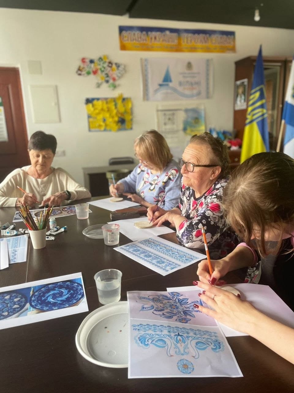 Чотири учасниці різного віку сидять у ряд за великим столом під час творчого заняття. Вони схилилися над столом, розписуючи дерев'яні основи олівцями та пензлями. На передньому плані дівчина у чорній кофті виводить візерунок, орієнтуючись на роздрукований паперовий ескіз із традиційними синьо-білими розписами та зображенням птахів. На світлій стіні позаду них висять патріотичні плакати, мапа України з кольорових долоньок та державний прапор.