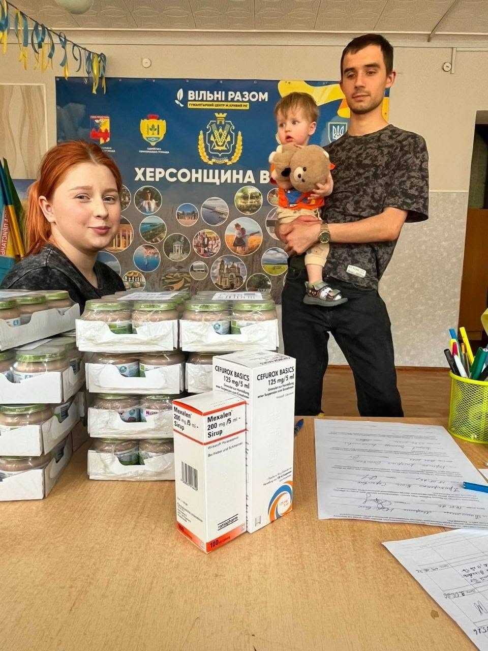 Видача допомоги в офіційному хабі. Ліворуч за столом стоїть рудоволоса дівчина, перед якою складені упаковки з дитячим харчуванням. Праворуч стоїть молодий чоловік у сірій камуфляжній футболці, який тримає на руках маленьку дитину з плюшевим ведмедиком. На передньому плані на столі крупно виділені дві біло-червоні картонні коробки з медичними сиропами «Mexalen» та «Cefurox Basics». Поруч лежать паперові бланки заяв та підставка з ручками. Позаду розміщено великий інформаційний банер гуманітарного центру.