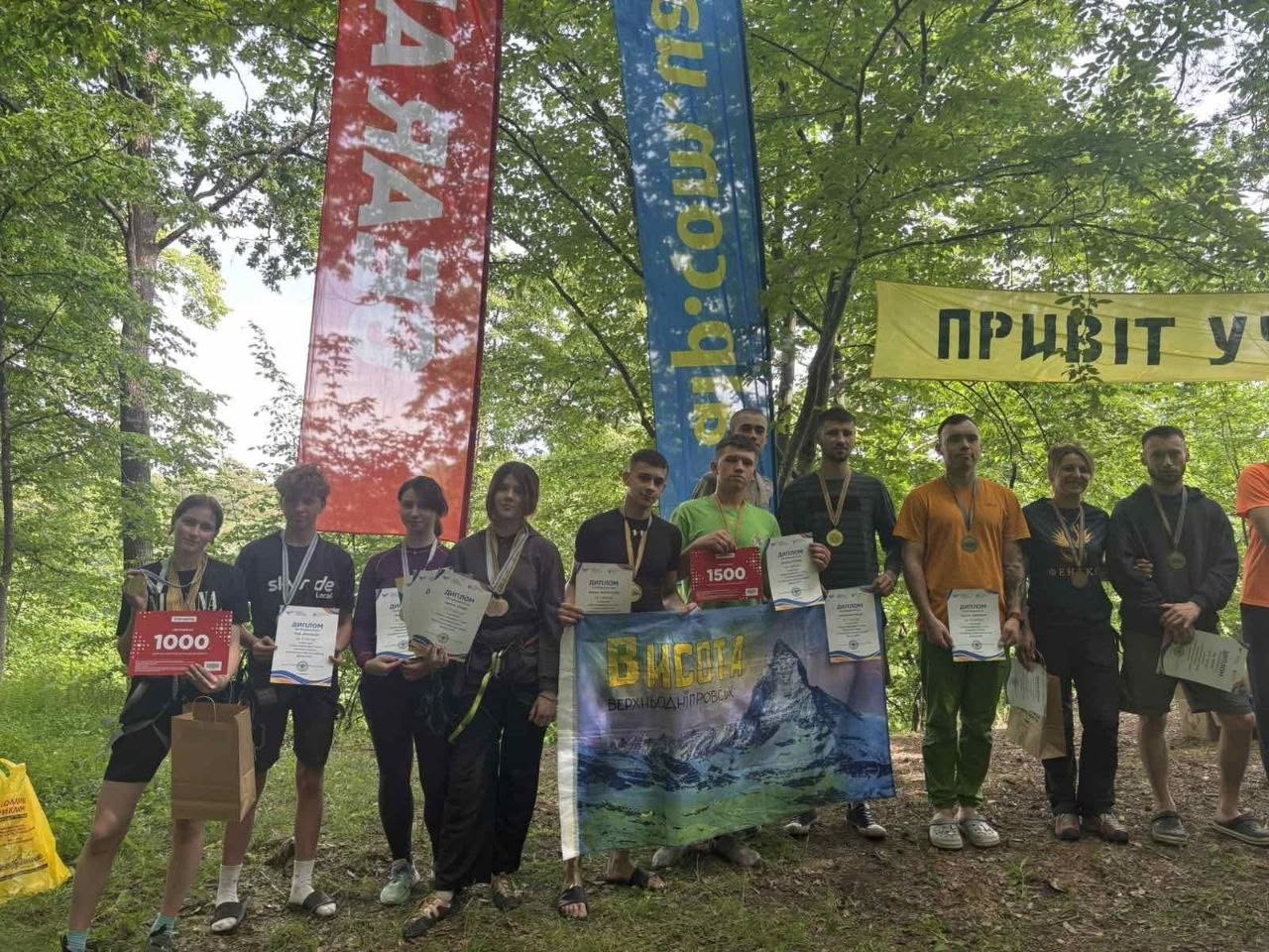 Фінальна церемонія нагородження учасників туристичного чемпіонату на лісовому майданчику. Одинадцятеро спортсменів (хлопці та дівчата) вишикувалися в один ряд. Вони тримають у руках почесні дипломи, медалі та призові сертифікати номіналом «1000» та «1500» гривень. Двоє хлопців по центру тримають розгорнутий прямокутний банер із зображенням засніженої гірської вершини та написом «ХЕРСОН. Верхньодніпровськ». Позаду учасників височіють великі червоні та блакитні прапори, а праворуч між деревами натягнуто жовту лінію з початком напису «ПРИВІТ У...».