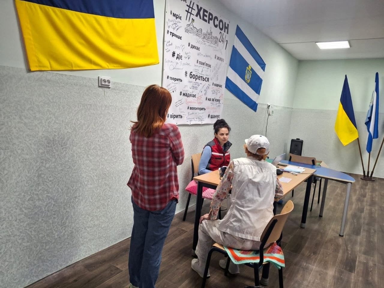 Консультативний прийом у світлому кабінеті з ламінатом на підлозі. За довгим дерев'яним столом сидить жінка-медик у синій кофті та червоному жилеті, яка розмовляє з відвідувачкою похилого віку у білій панамі та світлій блузці, що сидить навпроти на стільці. На передньому плані спиною до камери стоїть дівчина у червоній картатій сорочці та синіх джинсах. На стіні ліворуч розвішані державний прапор України, прапор міста Херсон та великий білий плакат із написом «#ХЕРСОН 24/7» і патріотичними хештегами з підписами від руки. На задньому плані праворуч стоять прапори України та ЄС.