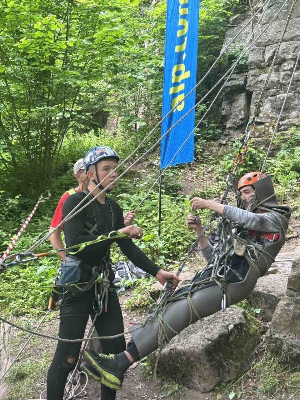 Практичне відпрацювання елементів порятунку та евакуації на гірському рельєфі. Справа зображено чоловіка в помаранчевій касці, який зафіксований у спеціальних армованих ношах-коконі у напіввертикальному положенні. Ноші підвішені на системі мотузок та карабінів. Ліворуч від нього стоїть молодий спортсмен у синій касці та чорному спортивному одязі зі страховочною системою, який обома руками регулює натяг мотузок. Заняття проходить у лісі біля підніжжя скелі на тлі великого вертикального блакитного прапора з жовтим написом.