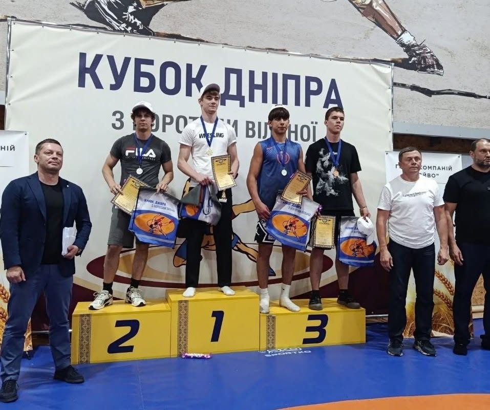 Церемонія нагородження юнаків на всеукраїнських змаганнях. Чотири спортсмени-призери стоять на жовтому п'єдесталі пошани на тлі великого білого банера з написом «КУБОК ДНІПРА З БОРОТЬБИ ВІЛЬНОЇ».  Переможець на першому місці вдягнений у синю спортивну кофту з тризубом, посміхається і піднімає вгору кубок.  Спортсмен на другому місці стоїть у сірому спортивному костюмі, тримаючи грамоту та синю сувенірну сумку.  На третьому місці стоять двоє борців: один у чорній футболці з медаллю на шиї, інший поруч із ним розгортає білий прапор Запорізької федерації вільної боротьби.  Ліворуч біля п'єдесталу стоїть чоловік у синьому піджаку, праворуч — чоловік у білій футболці. На передньому плані видно руки глядачів, один із яких знімає подію на смартфон.
