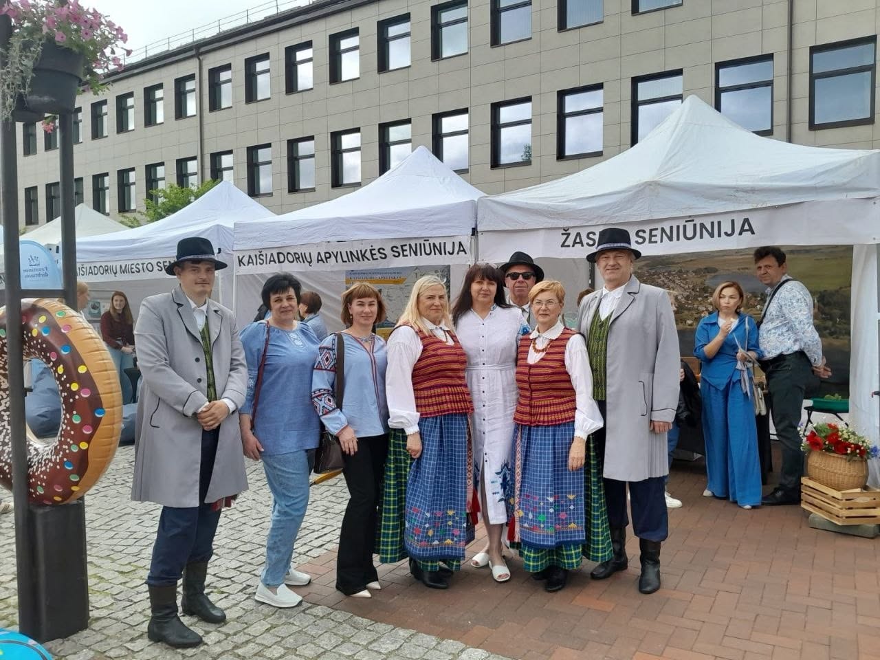 Учасники заходу на міській площі на тлі виставкових наметів. У центрі кадру стоять дев'ятеро людей, двоє з яких одягнені у традиційні литовські чи польські національні костюми (сірі пальта, капелюхи, смугасті спідниці), а частина українських представників — у блакитні та білі вишиванки. Ліворуч на передньому плані видно частину декоративного надувного пончика. Позаду групи розміщені білі ярмаркові намети з написами литовською мовою, зокрема «ŽASLIŲ SENIŪNIJA» та «KAIŠIADORIŲ APYLINKIŲ SENIŪNIJA». На задньому плані підноситься багатоповерхова адміністративна будівля.