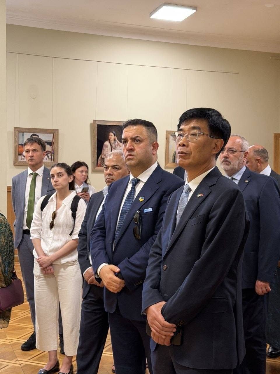 Члени офіційної міжнародної делегації крупним планом під час екскурсії. На передньому плані праворуч стоїть азійський дипломат в окулярах, темному костюмі та світло-блакитній краватці, на лацкані його піджака прикріплено маленький прапорець. Поруч із ним стоїть інший представник делегації у темно-синьому костюмі з жовто-блакитним значком на лацкані. На другому плані ліворуч видно дівчину в білій сорочці, чоловіка в сірому піджаку з зеленою краваткою та інших учасників заходу, які зосереджено дивляться ліворуч в один бік. На задньому плані на стіні розмито видно картини.