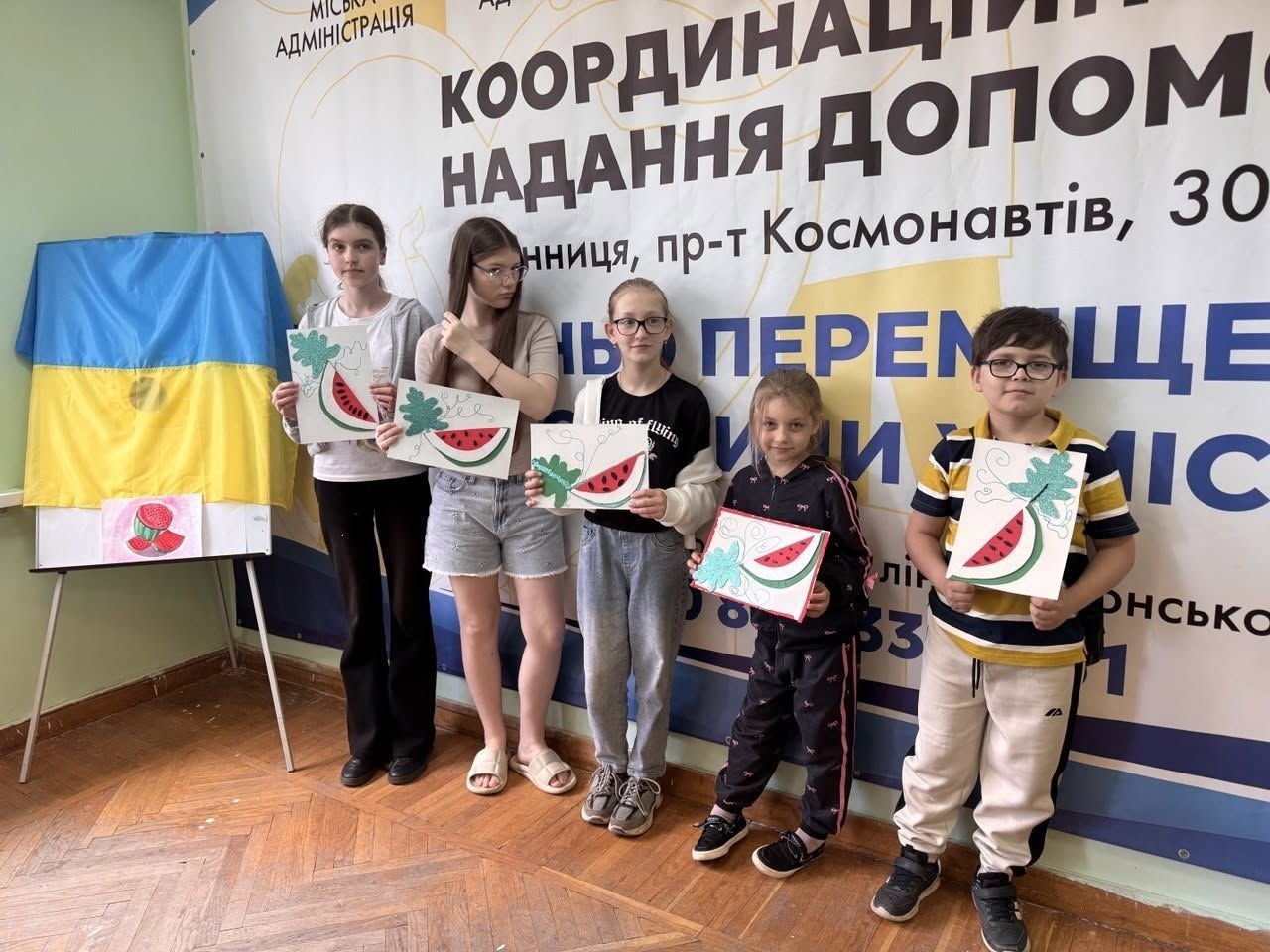 Загальний план творчої майстерні для дітей та дорослих у координаційному хабі. Навколо довгого великого дерев'яного столу сидять близько десяти учасників заходу, які займаються рукоділлям, вирізаючи та склеюючи паперові елементи. На столі розкладено кольоровий папір, ножиці, олівці, а в центрі стоїть велика купа різнокольорових пластикових деталей та конструктора. Наприкінці столу стоїть жінка-інструктор у зеленій сорочці, яка керує процесом. Позаду неї на стіні закріплено офіційний великий банер Координаційного центру допомоги ВПО з Херсонщини у місті Вінниця, на якому зверху зображено логотипи Вінницької, Херсонської, Генічеської та Ювілейної адміністрацій. Справа на столі видно монітор комп'ютера та принтер.