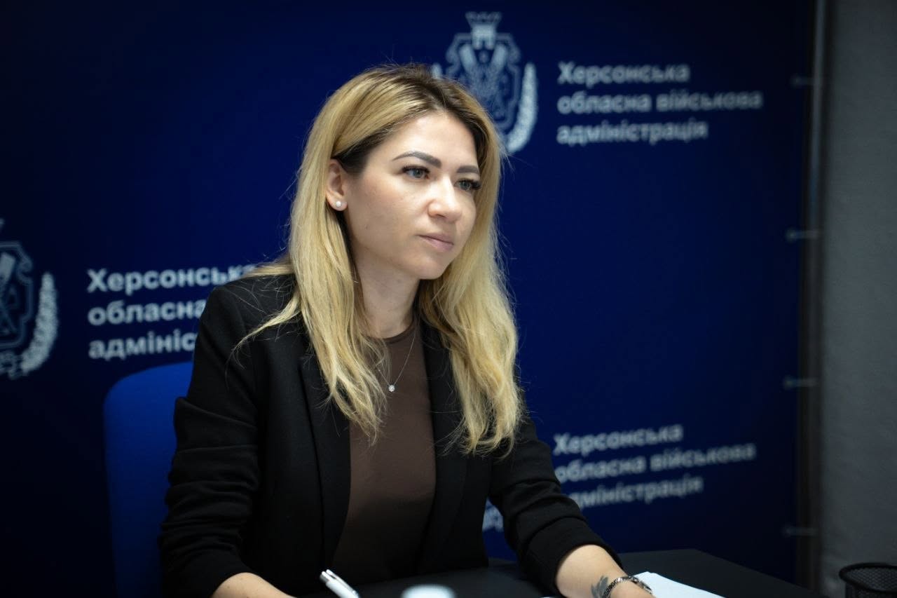 Портретне фото заступниці начальника Херсонської обласної військової адміністрації Ольги Малярчук під час наради. Жінка зі світлим волоссям зображена у напівпрофіль, вона одягнена в чорний піджак, коричневий топ і тонку підвіску на шиї. Вона зосереджено дивиться вбік, сидить за робочим столом, тримаючи в руці білу ручку.  Позаду неї розташовано суцільний темно-синій пресволл (банер) із повторюваним білим логотипом (гербом області) та офіційним написом «Херсонська обласна військова адміністрація».