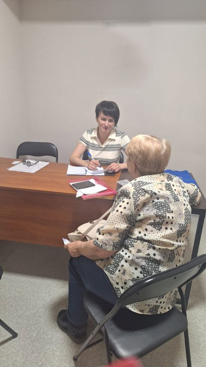 Індивідуальна юридична або адміністративна консультація в кабінеті. За великим дерев'яним столом коричневого кольору сидить молода жінка-спеціаліст із короткою темною зачіскою, одягнена у білу футболку в тонку чорну смужку. Вона уважно слухає відвідувачку, тримаючи ручку над розгорнутим блокнотом.  Навпроти неї спиною до камери на чорному розкладному стільці сидить літня жінка у світлій блузці з дрібним візерунком та синіх джинсах, яка тримає в руці невеликий папірець. На її плечі висить тонка світла сумочка. На столі лежать документи, канцелярське приладдя та мобільний телефон.