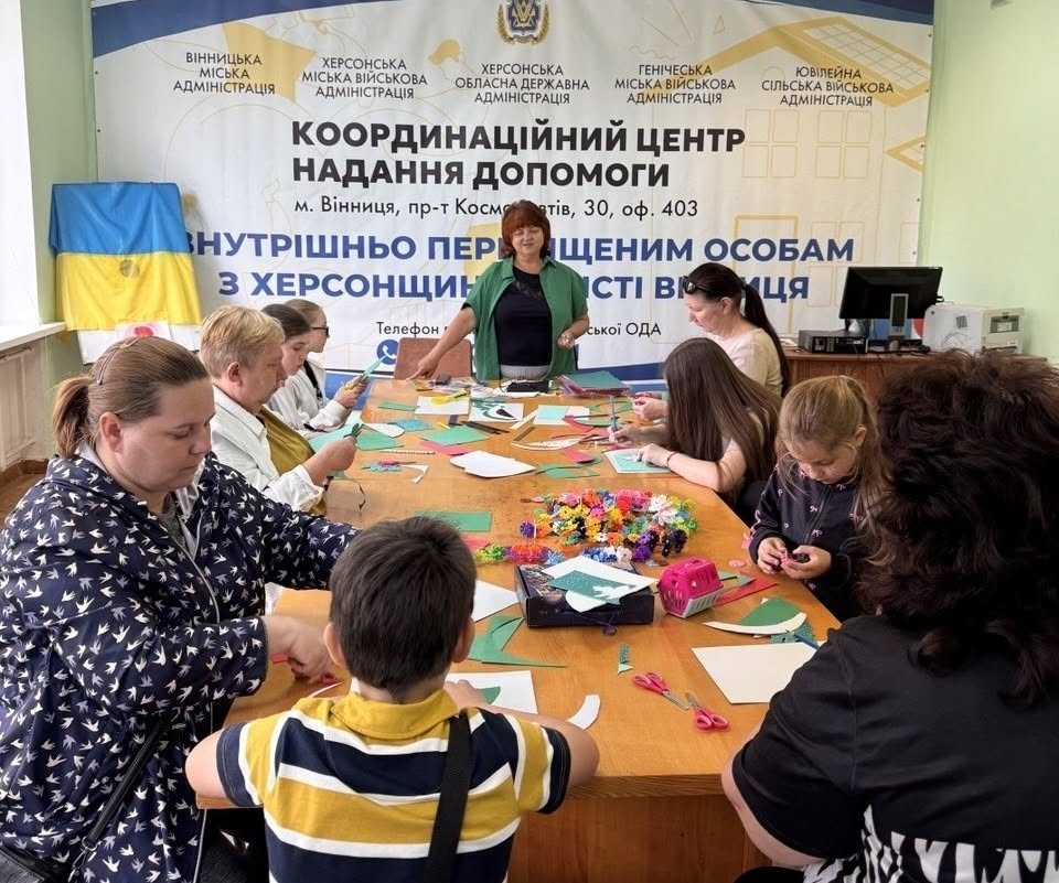 П'ятеро дітей різного віку (чотири дівчинки та хлопчик) стоять в один ряд у приміщенні хабу, тримаючи в руках свої готові творчі роботи. На аркушах паперу виконано об'ємні аплікації у вигляді часточки кавуна із зеленим хвостиком. Діти одягнені в літній повсякденний одяг. Позаду них на стіні розміщено великий білий банер із синім та чорним текстом: «КООРДИНАЦІЙНИЙ ЦЕНТР НАДАННЯ ДОПОМОГИ... м. Вінниця, пр-т Космонавтів, 30... ВНУТРІШНЬО ПЕРЕМІЩЕНИМ ОСОБАМ З ХЕРСОНЩИНИ». Ліворуч на маленькому мольберті встановлено державний прапор України, під яким стоїть ще один дитячий малюнок кавуна.