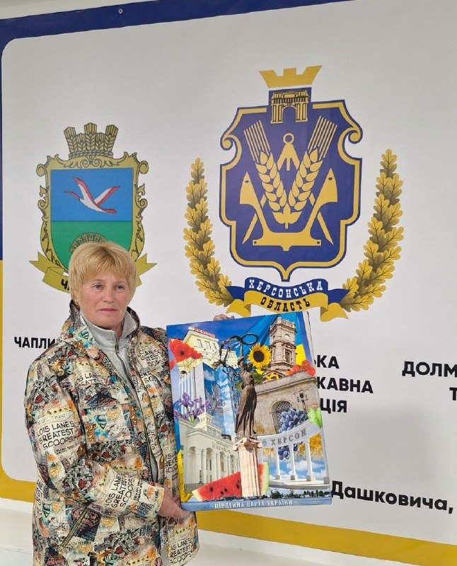 Жінка у світлій куртці з газетним принтом тримає в руках плакат із колажем про Херсон. Вона стоїть на тлі білої стіни, де зображені великі герби: праворуч — герб Херсонської області (із синім щитом, якорем та колоссям), ліворуч — герб селища Чаплинка.
