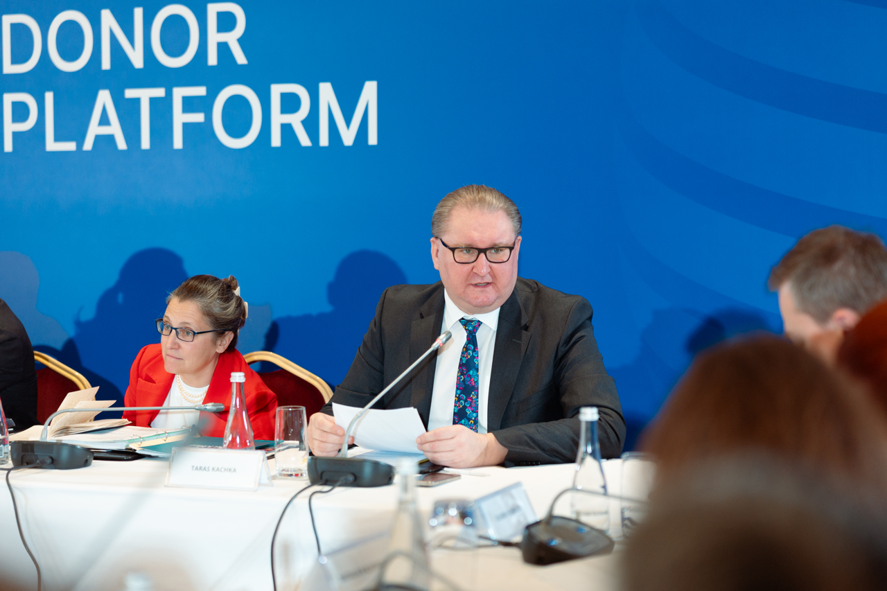 Чоловік у діловому костюмі та окулярах виступає з промовою, тримаючи в руках аркуш паперу. Поруч із ним сидить жінка в червоному піджаку, яка уважно слухає. Вони перебувають за столом засідань на фоні великого синього банера з написом «DONOR PLATFORM».