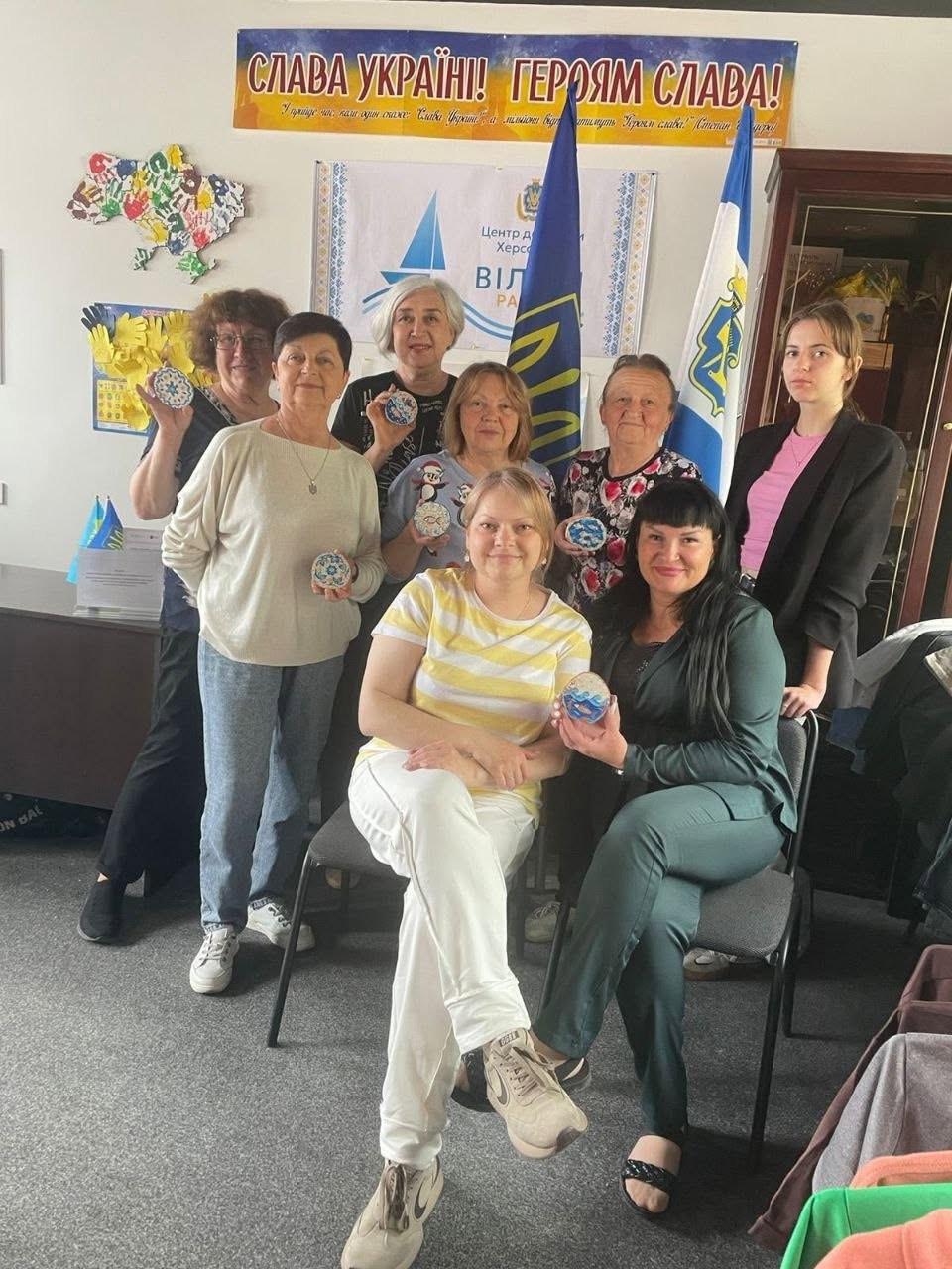 Спільне групове фото учасниць та організаторів майстер-класу в приміщенні хабу. Вісім жінок позують перед камерою, тримаючи в руках розписані синьо-білими орнаментами круглі дерев'яні сувеніри. Дві жінки сидять на стільцях на передньому плані, інші шість стоять за ними у повний зріст. На задньому плані встановлено державний прапор України, прапор міста Херсон та великий жовто-блакитний банер із гаслом «СЛАВА УКРАЇНІ! ГЕРОЯМ СЛАВА!».