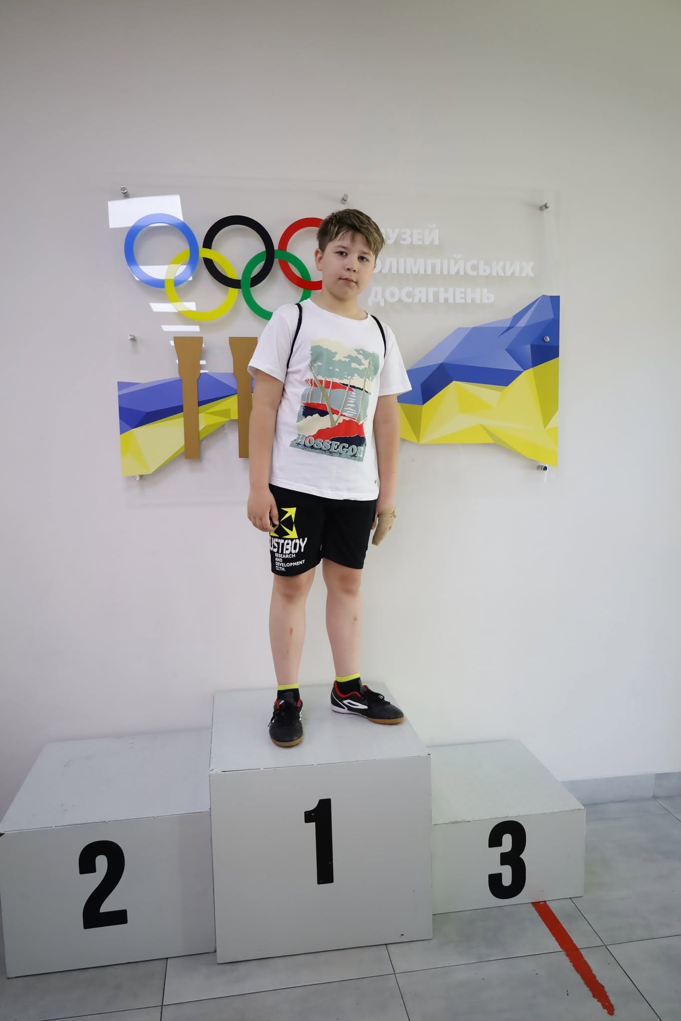 Хлопчик шкільного віку стоїть у повний зріст на білому дерев'яному п'єдесталі пошани, зайнявши перше місце (цифра 1). Він одягнений у білу футболку з літнім принтом та написом «HOSSEGOR», чорні спортивні шорти з білим текстом «JUSTBOY» та чорні кросівки з червоними вставками. Його ліва кисть обмотана еластичним бинтом. Хлопчик позує на тлі чистої білої стіни, де закріплено прозорий пластиковий стенд із різнокольоровими олімпійськими кільцями, жовто-блакитним шлейфом у вигляді прапора України та написом: «Музей олімпійських досягнень».
