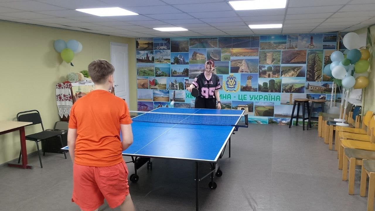 Двоє підлітків грають у настільний теніс (пінг-понг) у просторій ігровій кімнаті. На передньому плані спиною до камери стоїть хлопець у яскравій помаранчевій футболці та червоних шортах. Навпроти нього за синім тенісним столом із натягнутою сіткою стоїть дівчина в чорній спортивній кофті з цифрою «95», яка готується відбити ракеткою білу кульку. Дальня стіна зали повністю задекорована великим фотоколажем із краєвидами міст та пам'яток, поверх якого нанесено напис «...МИНА — ЦЕ УКРАЇНА». По кутах кімнати розвішані зв'язки блакитних, жовтих та зелених надувних кульок, а праворуч стоять дерев'яні стільці.